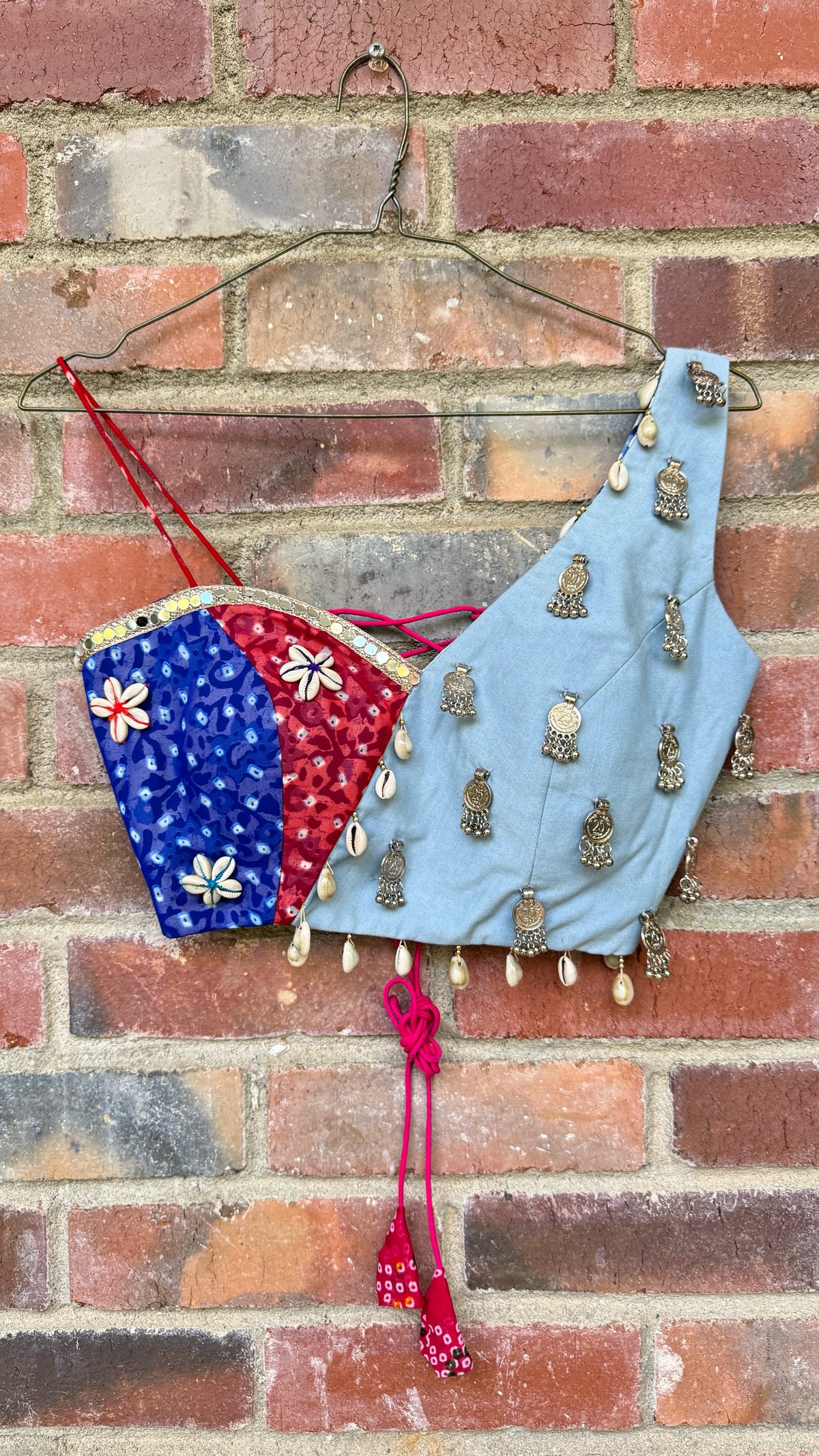 A light blue sleeveless blouse top with metallic coin embellishments and a red tassel, shown hanging on a wire hanger against a red brick wall. 
